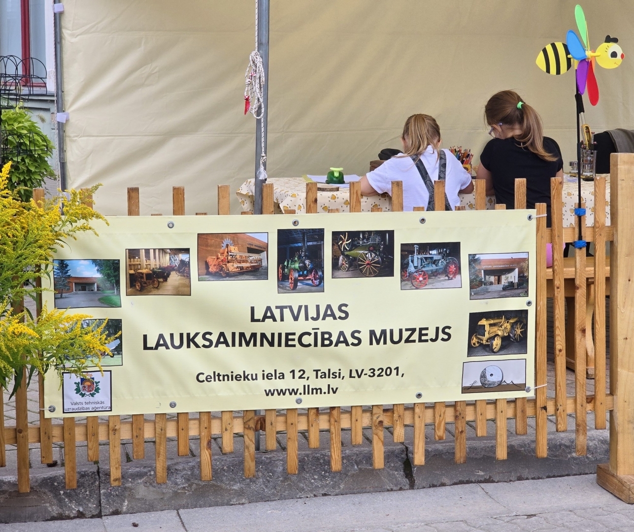 Latvijas Lauksaimniecības muzeja baneris uz gaišas koka sētas. Fonā divas meitenes un vēja ķērājs bites formā