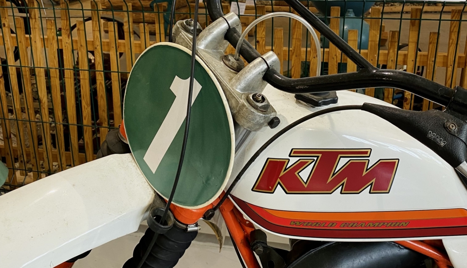 Krosa motocikla KTM stūres un bākas daļa. Motocikla numurs viens. Fonā muzeja kāda no izstādes sienām